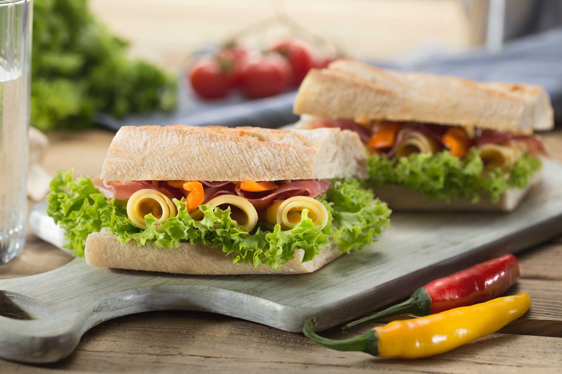 Two pieces of sandwich with thick bread, lettuce, cheese, meat, and something orange that is not cheese. A chili in the foreground and a glass of water partially in frame.