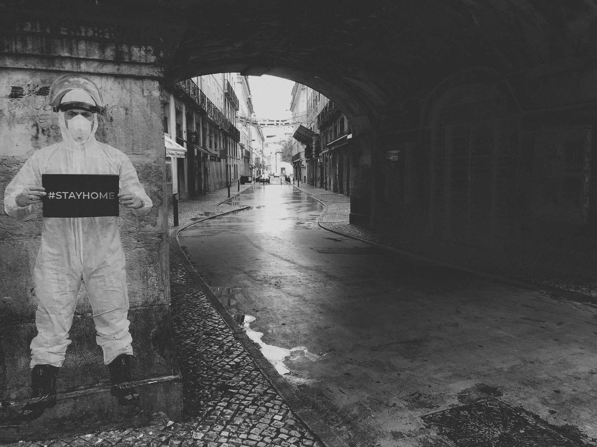 Worker in hazmat gear near an empty street holding a sign that says "#STAYHOME"