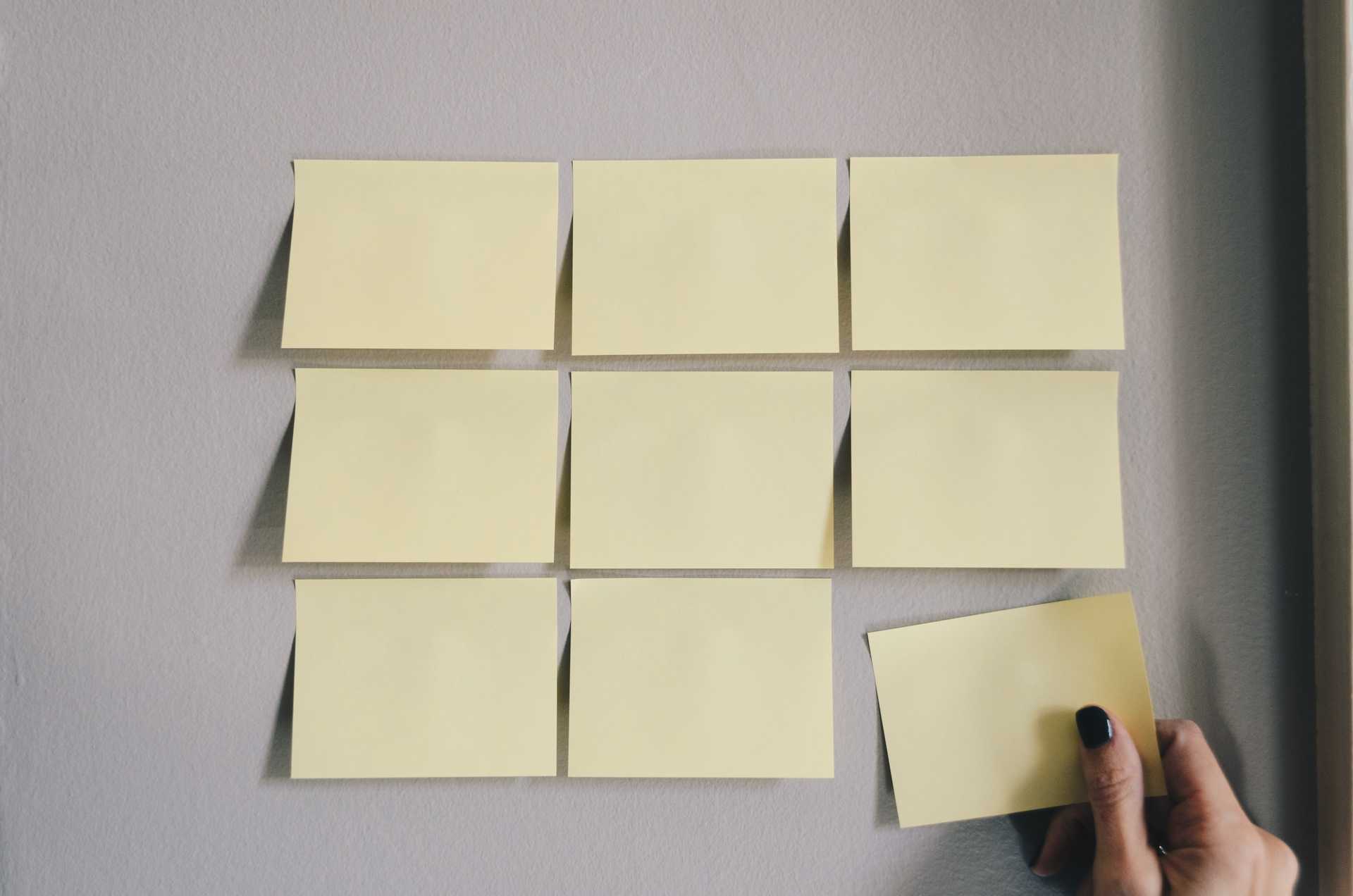 A 3 by 3 matrix of yellow post-it notes on a white background with the last being placed by a barely seen hand