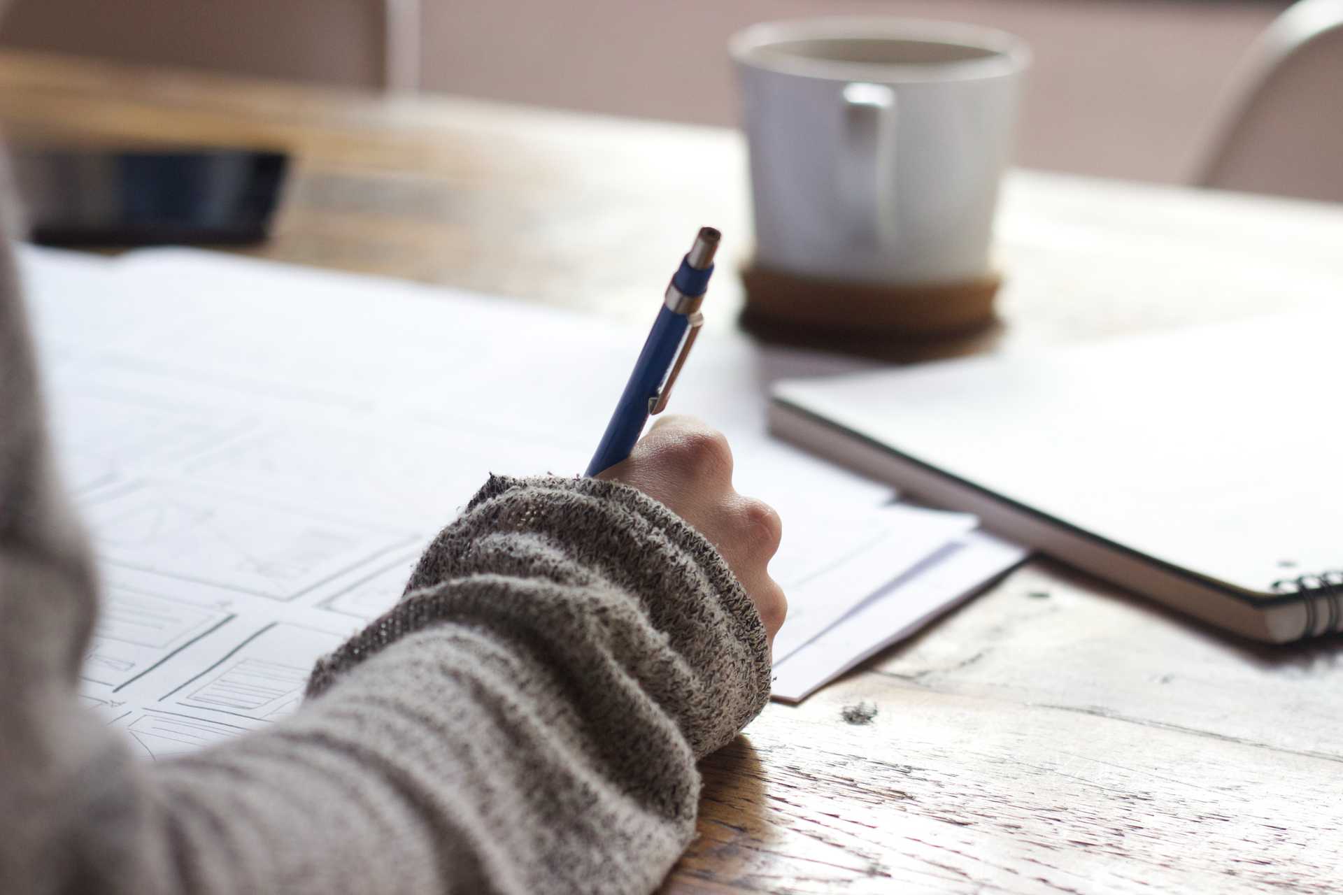 A person writing with a blue mechanical pencil wearing a long sleeve light brown and gray shirt or sweater. Only a bit of sleeve is visible. A notebook and coffee mug are in the background.