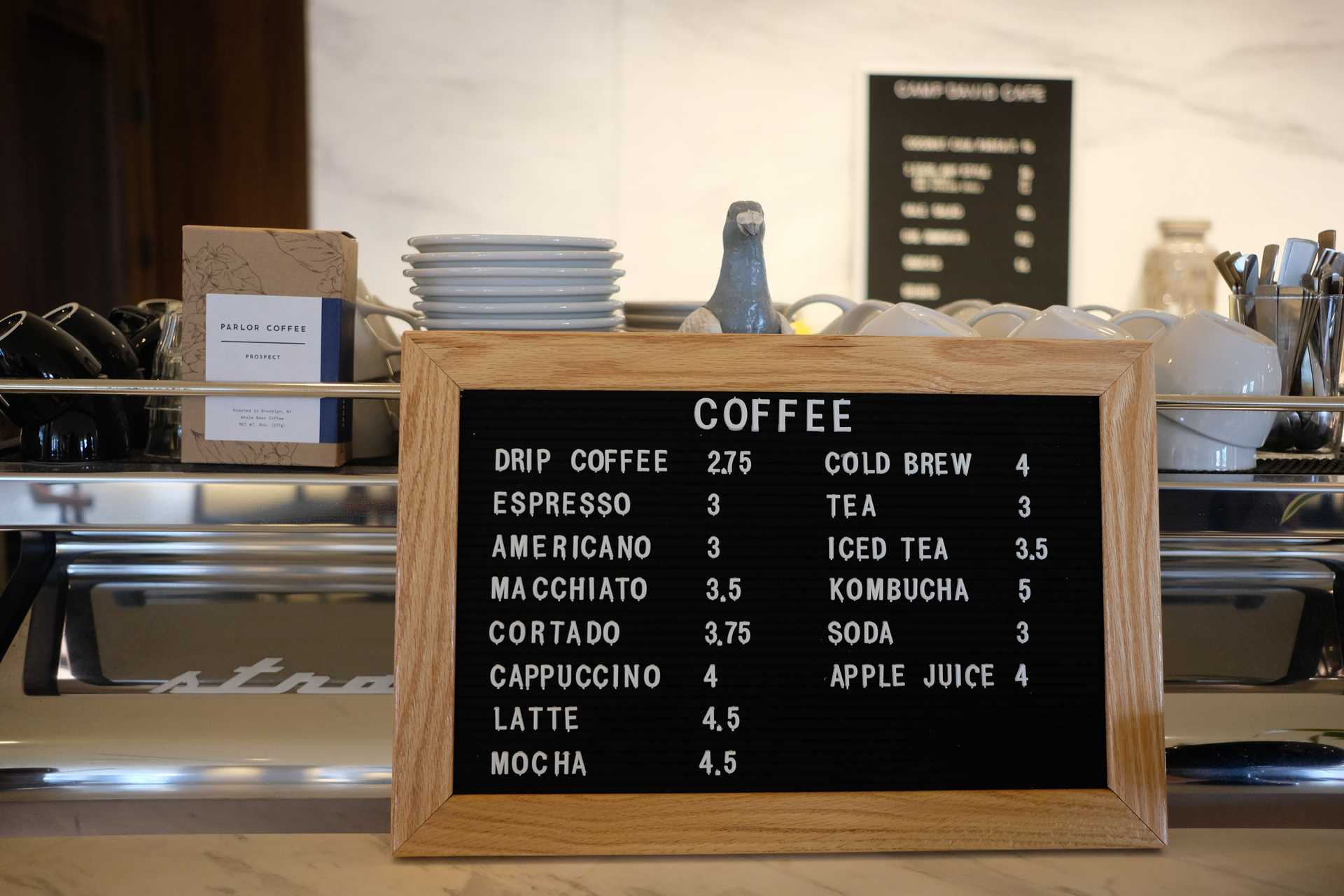 Menu at a coffee shop displaying various drinks and their price. In the background, there is a statue of a pigeon looking at us.